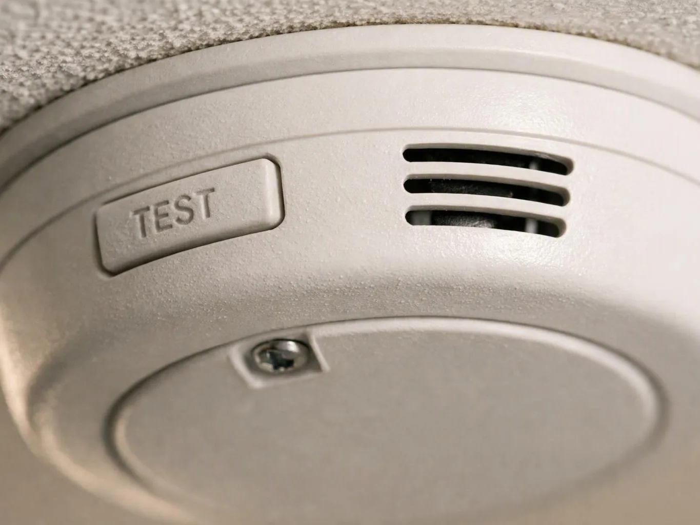 Close-up of a ceiling smoke detector with a TEST button and vent slots on a textured ceiling