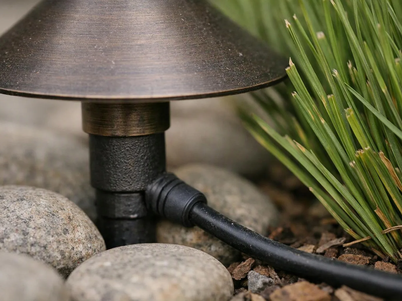 A garden light sits among smooth stones and pine needles, with a black cable running beside green blades